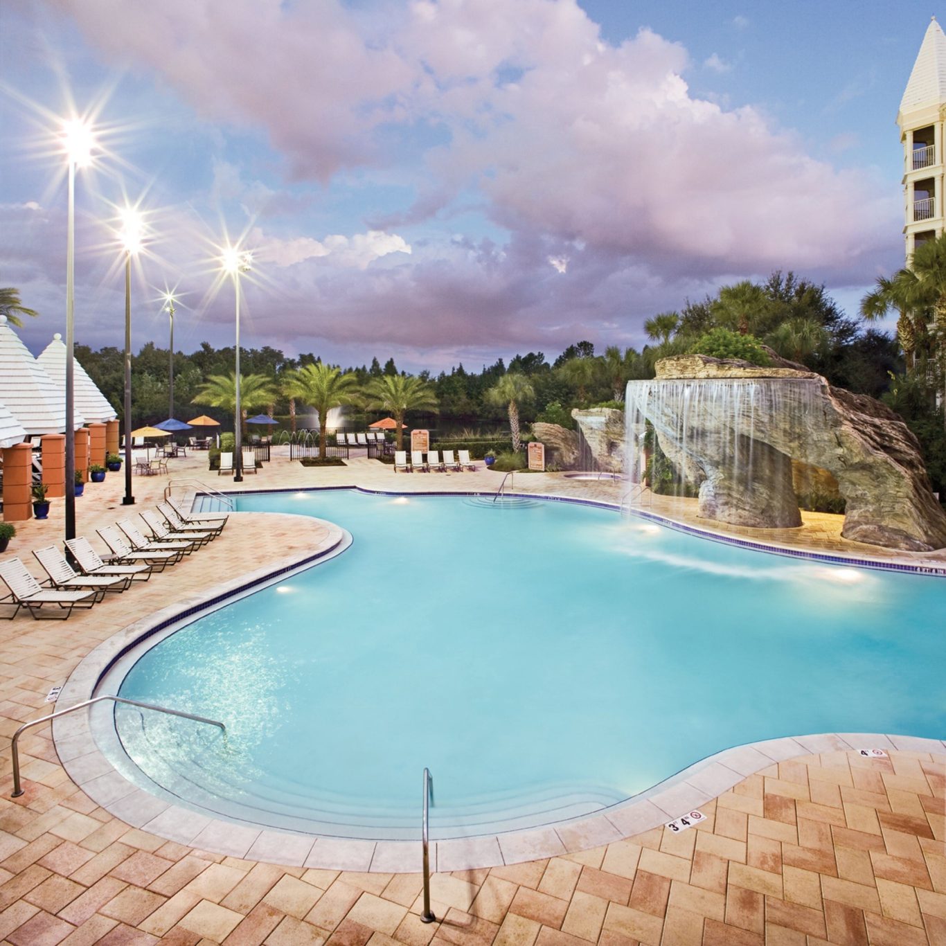 Swimming pool with a waterfall, lounge chairs, and palm trees under a cloudy sky.