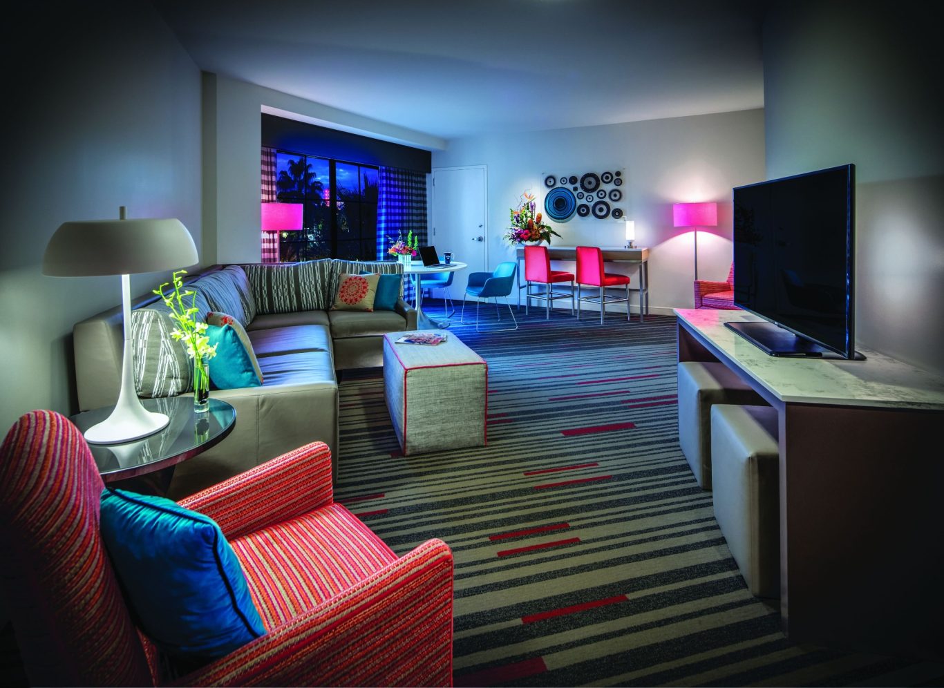 Modern hotel room featuring a sofa, armchair, TV, and colourful accent lighting.
