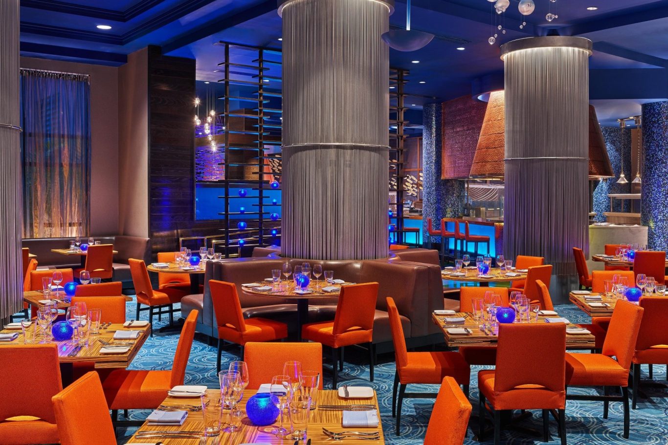 Modern restaurant interior with blue lighting and orange chairs, set for dining.