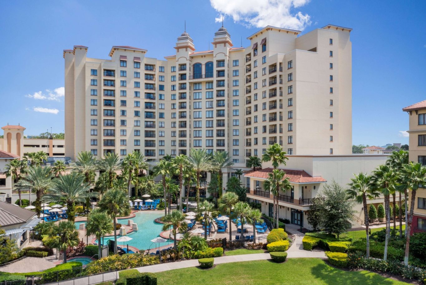 A luxurious hotel with palm trees and a large swimming pool in a sunny setting.
