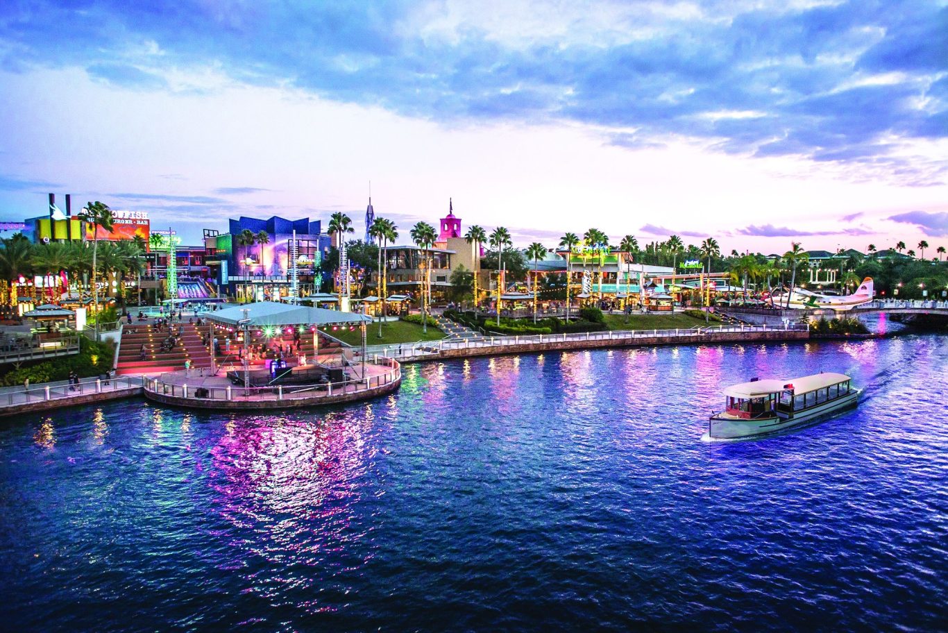Colourful waterfront scene with buildings, palm trees, and a boat on the water at sunset.