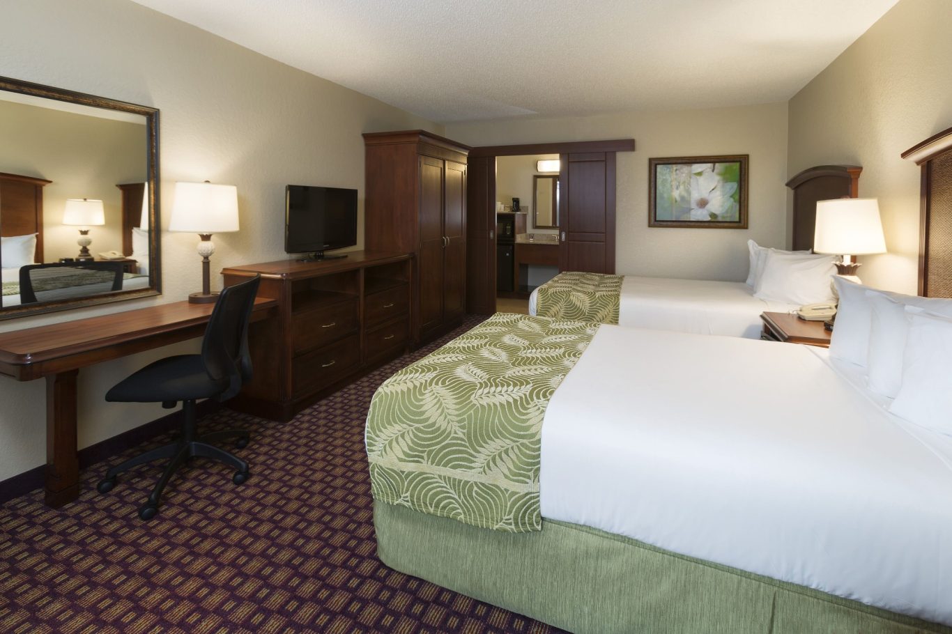 Hotel room with two beds, a desk, television, and warm lighting.