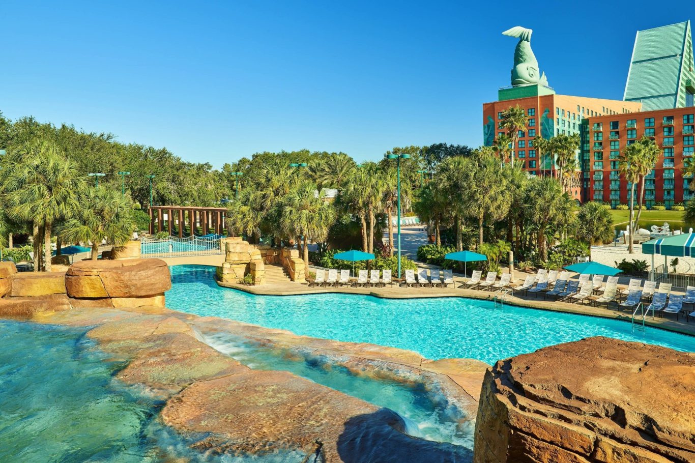 Swimming pool surrounded by palm trees and rocks, with a hotel in the background.