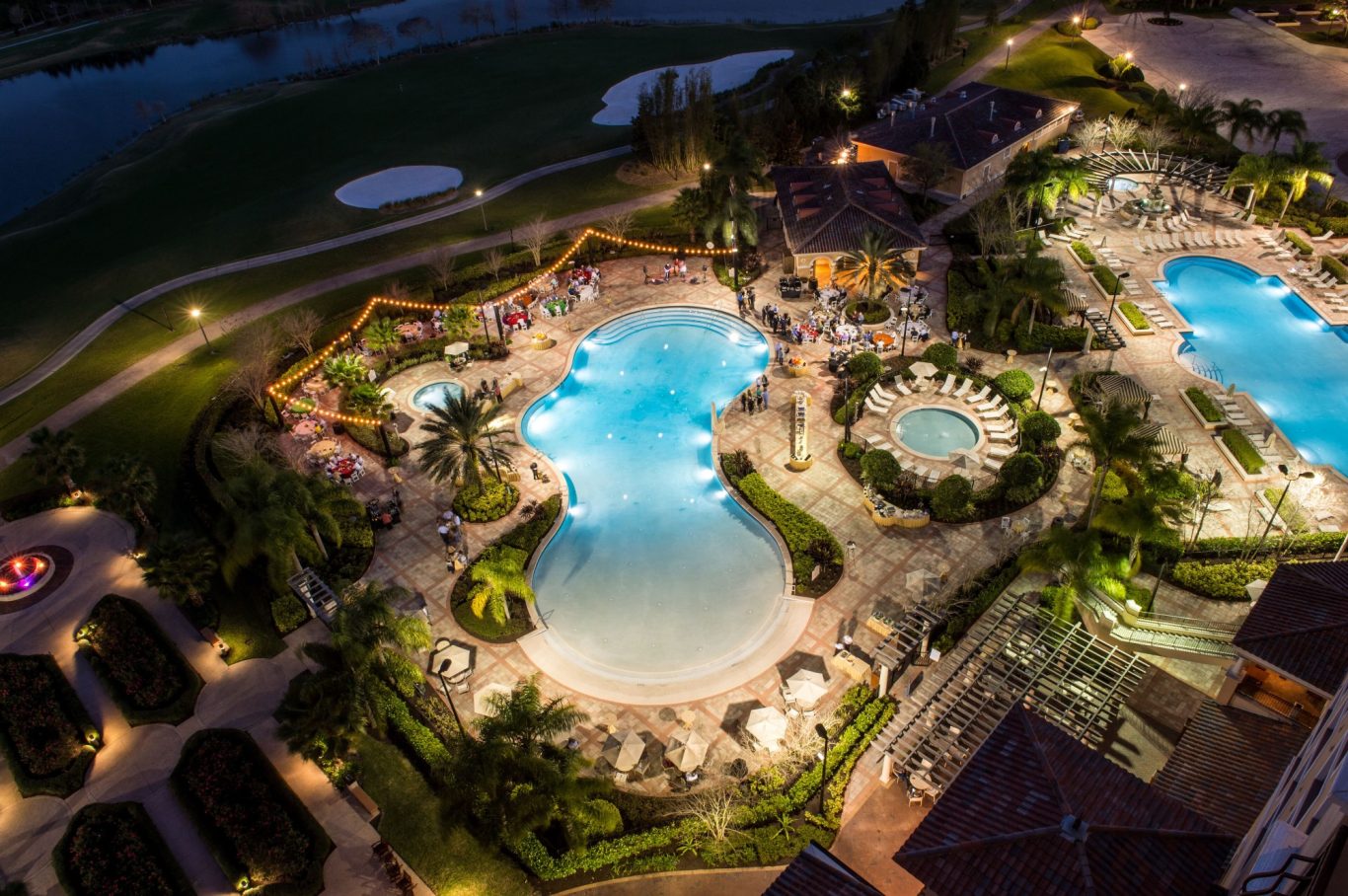 Aerial view of a resort pool area surrounded by palm trees and lounge chairs at night.
