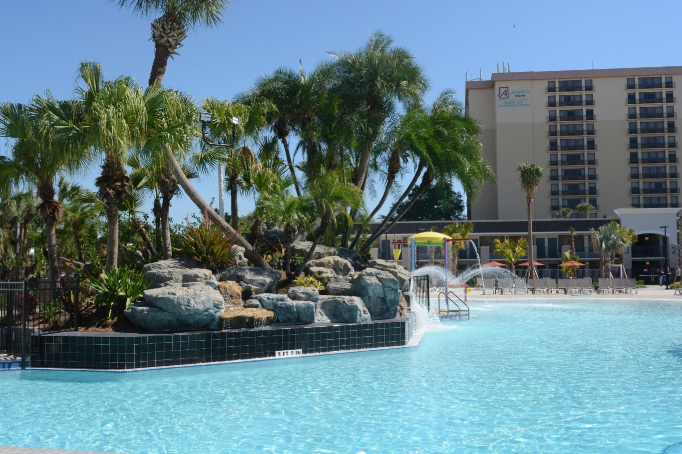 A large pool surrounded by palm trees and a hotel in sunny weather.