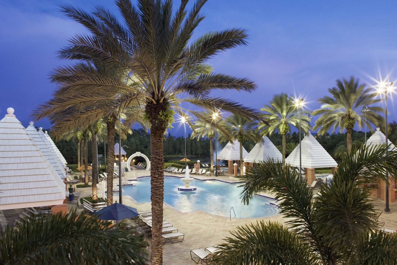 Tropical resort with palm trees, a pool, and iconic white tent-like structures at dusk.