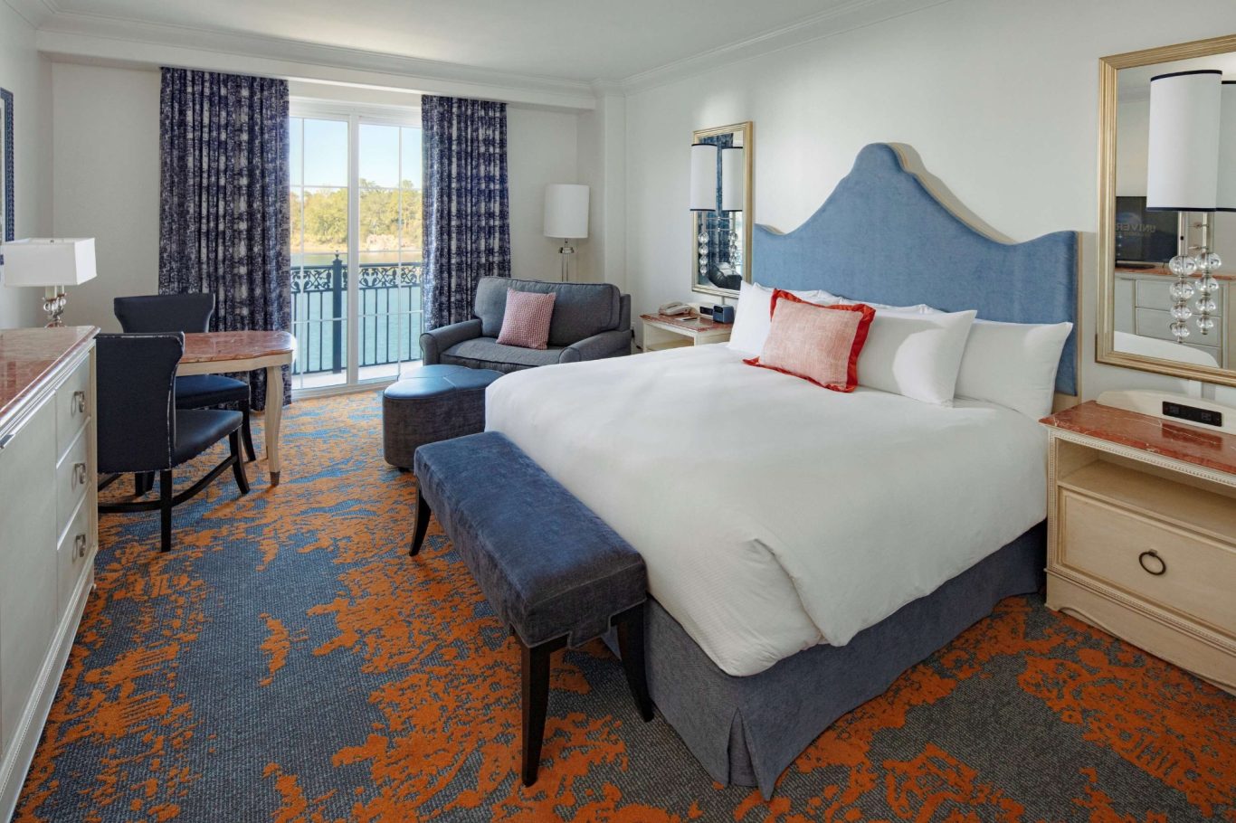 A stylish hotel room featuring a queen bed, seating area, desk, and large window.