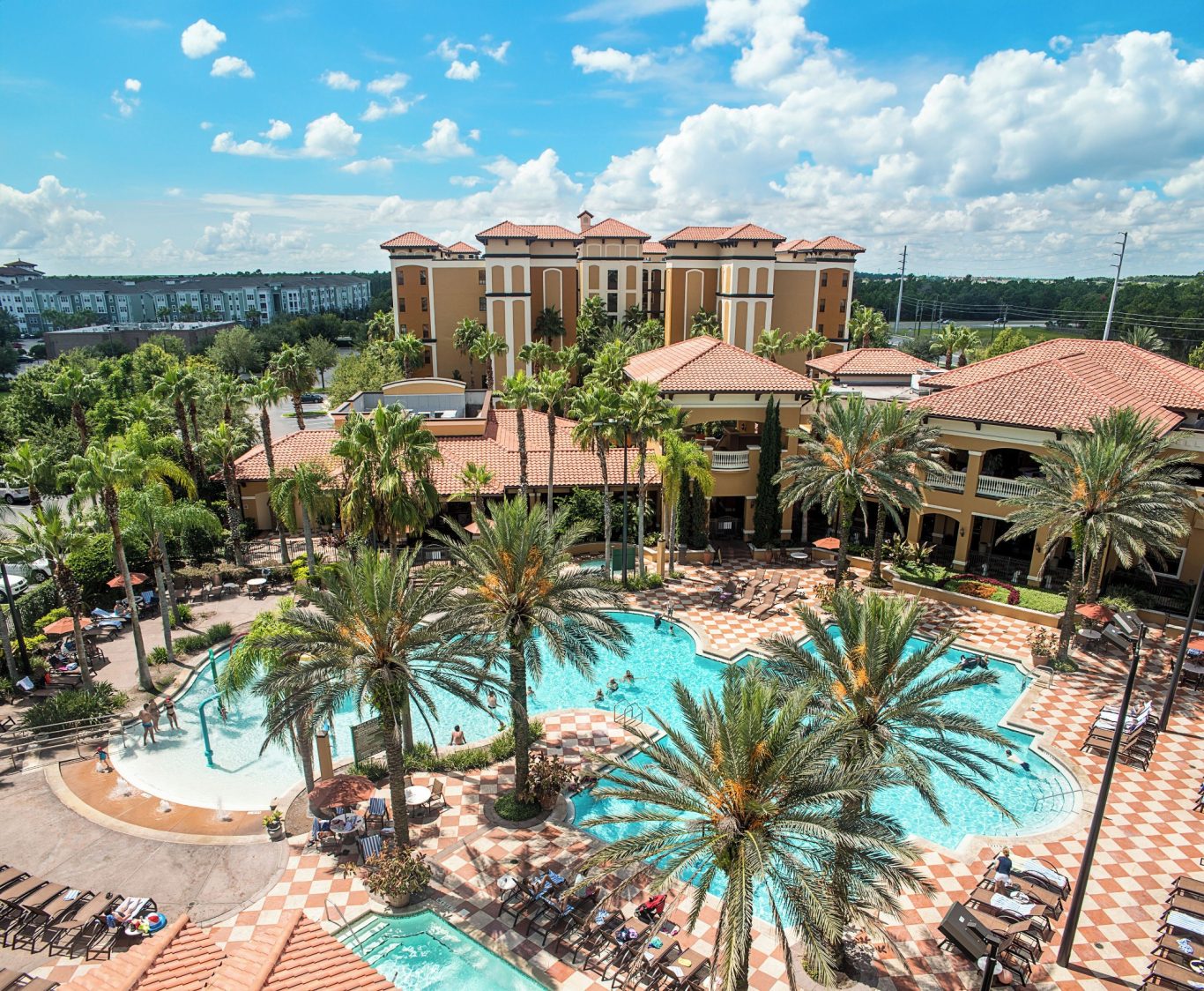 Aerial view of a hotel with a pool surrounded by palm trees and blue skies.