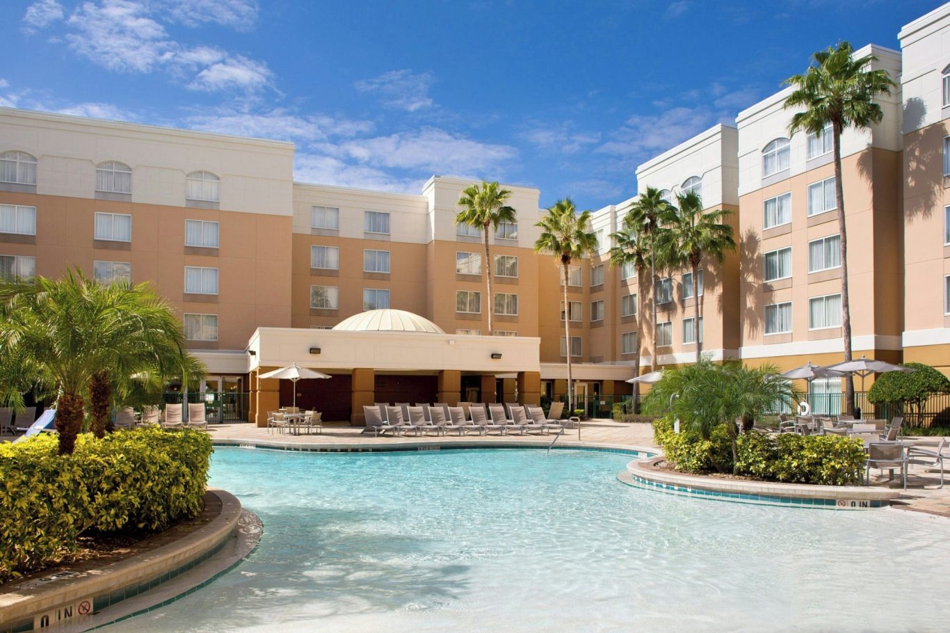 Swimming pool surrounded by palm trees and a hotel building against a blue sky.