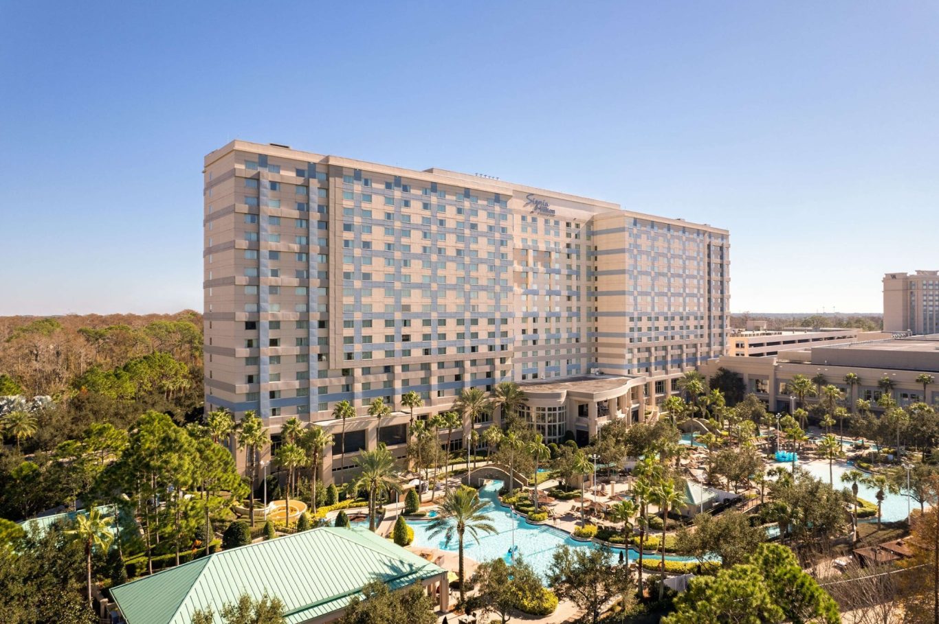 A modern hotel building surrounded by greenery and a pool area under a clear blue sky.