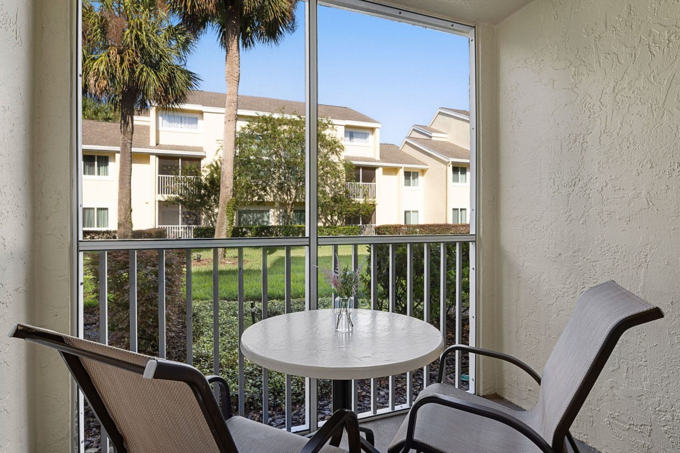 A small balcony with a round table and chairs, overlooking a green courtyard and buildings.