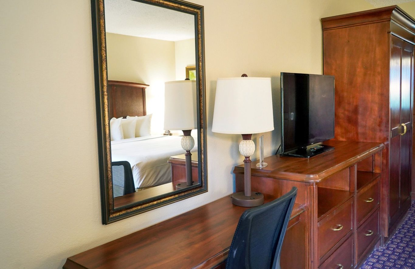 Hotel room interior with a desk, lamp, mirror, and television.
