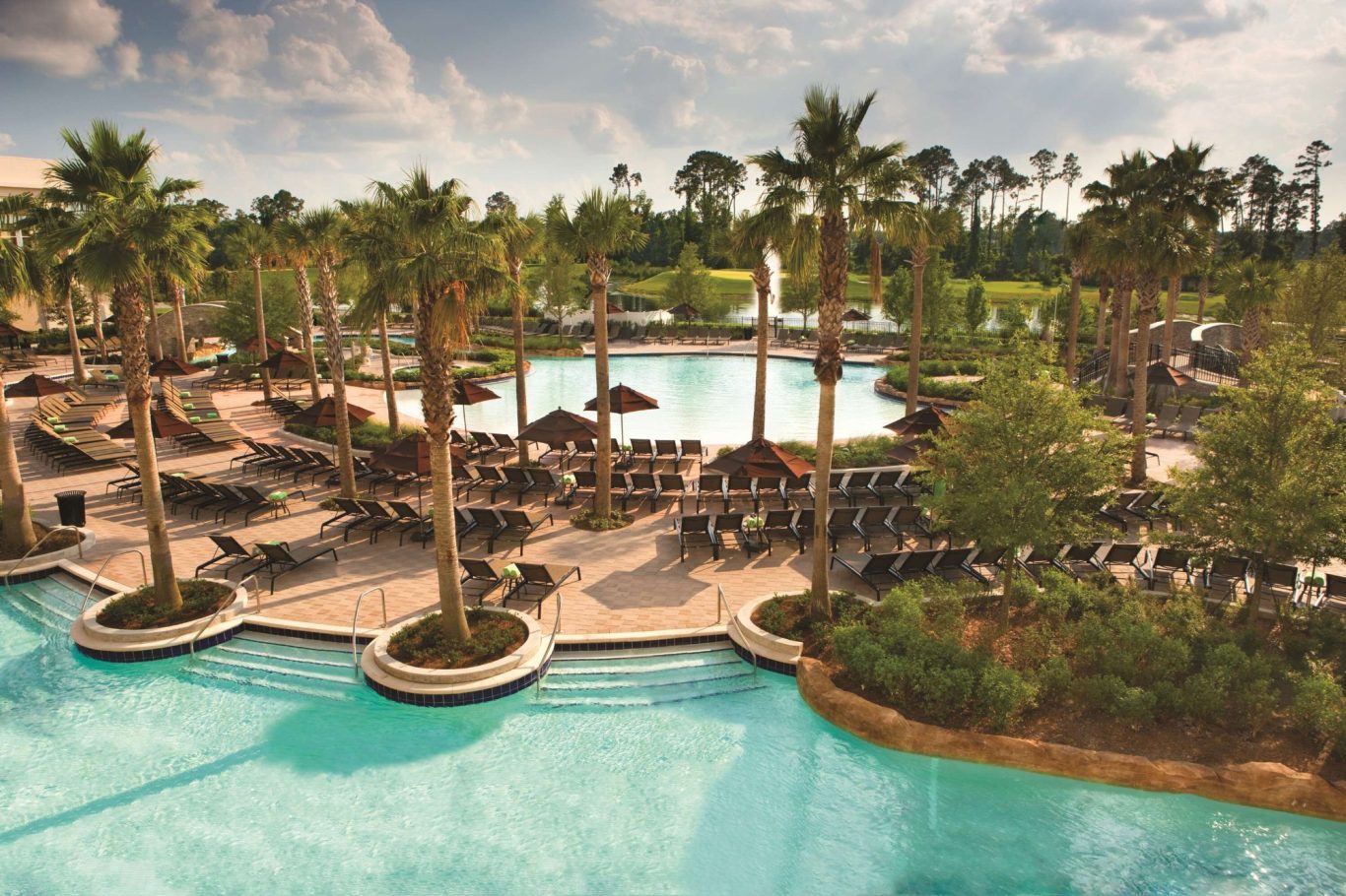 A resort pool area surrounded by palm trees and lounge chairs under a cloudy sky.