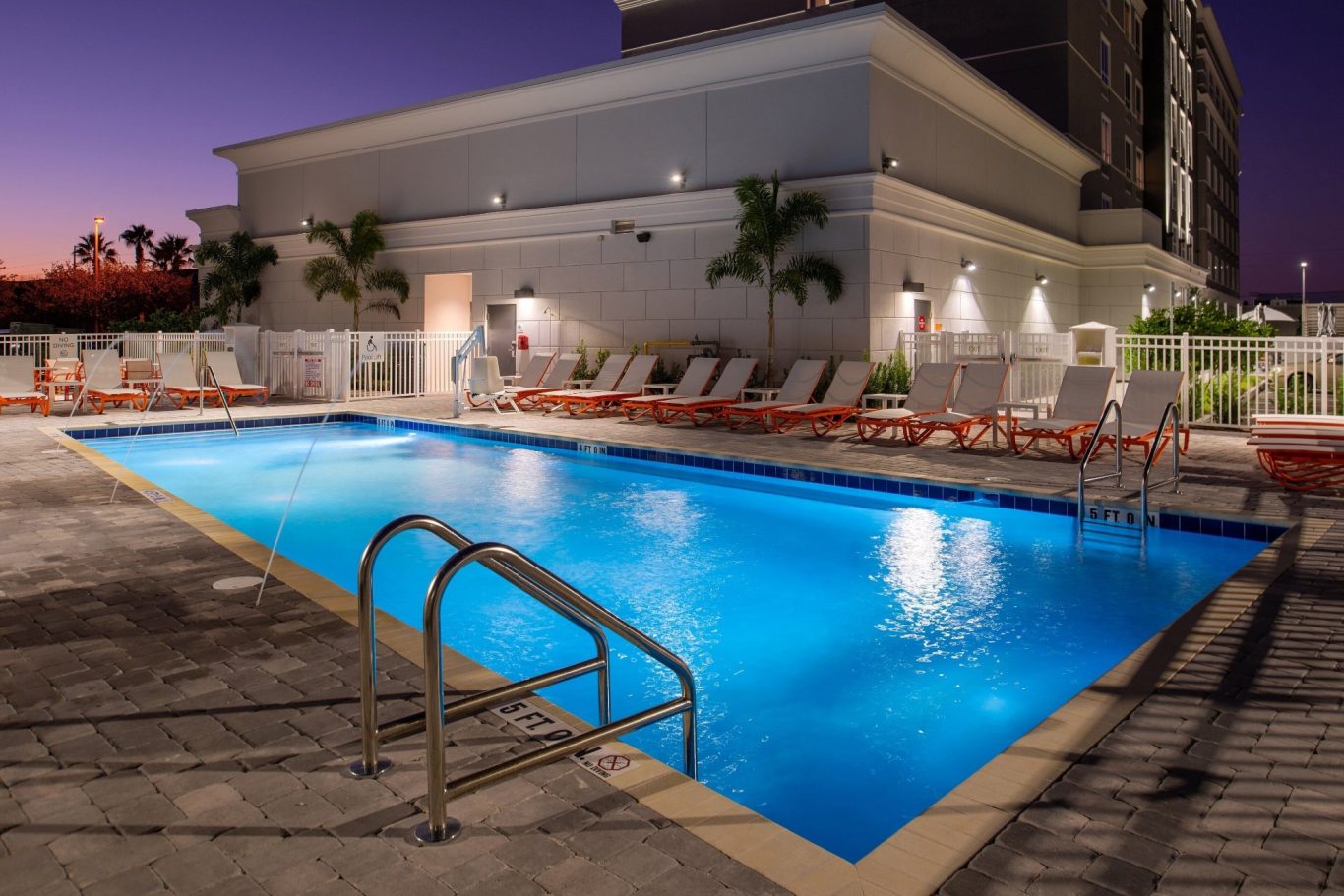 A well-lit swimming pool surrounded by lounge chairs at dusk.