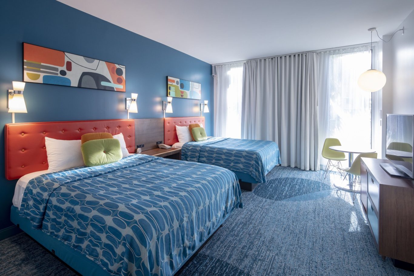 Modern hotel room with two beds, bright blue walls, and a stylish workspace.