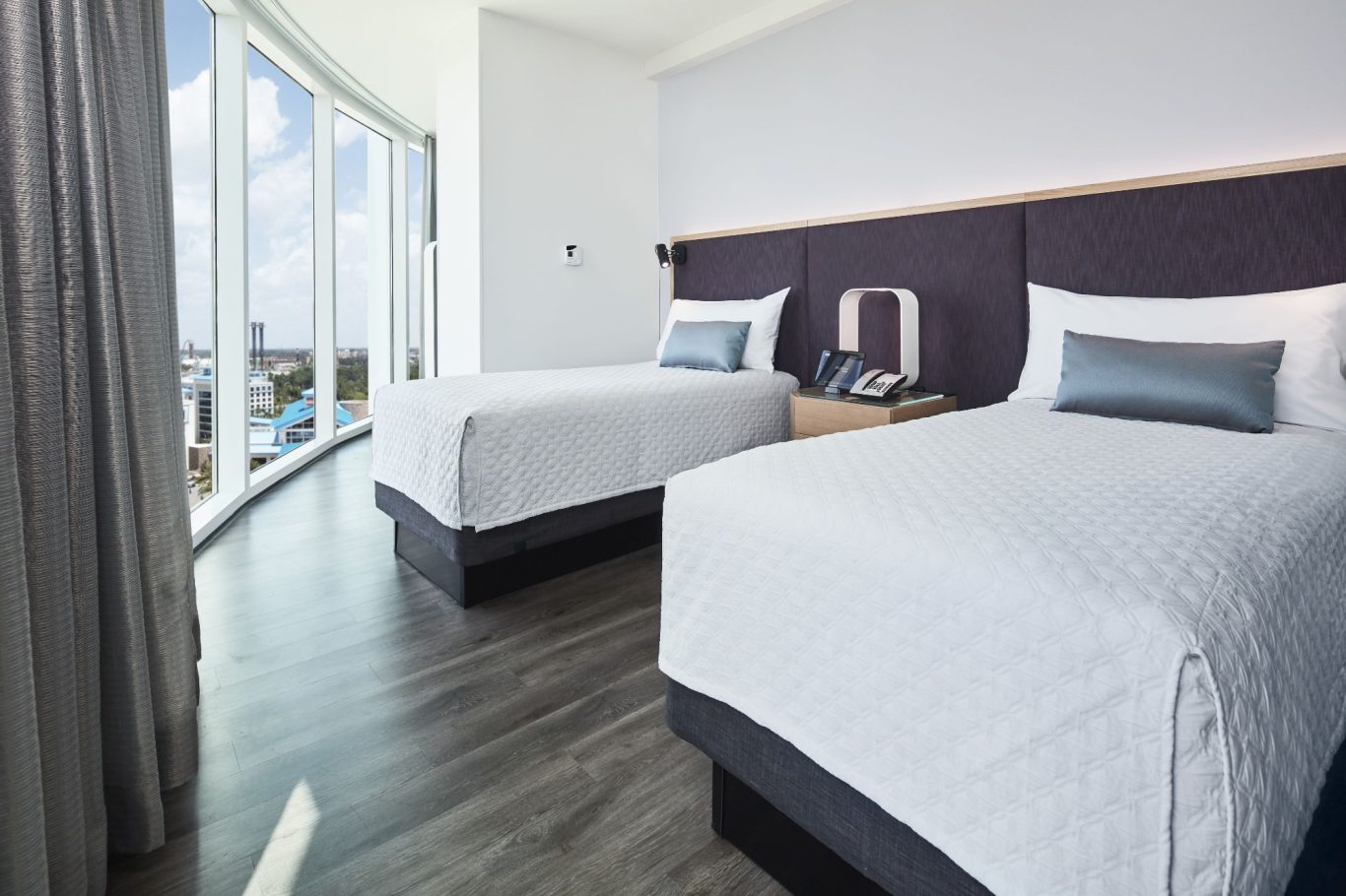 Modern hotel room with two beds, large windows, and contemporary furnishings.