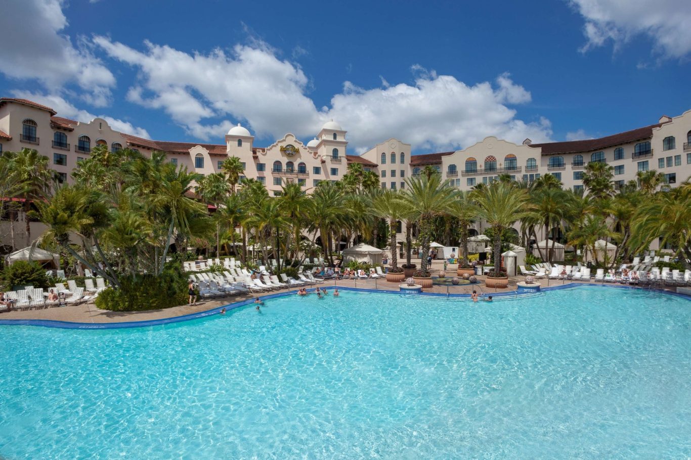 Clear blue pool surrounded by palm trees and a hotel with sunny skies.