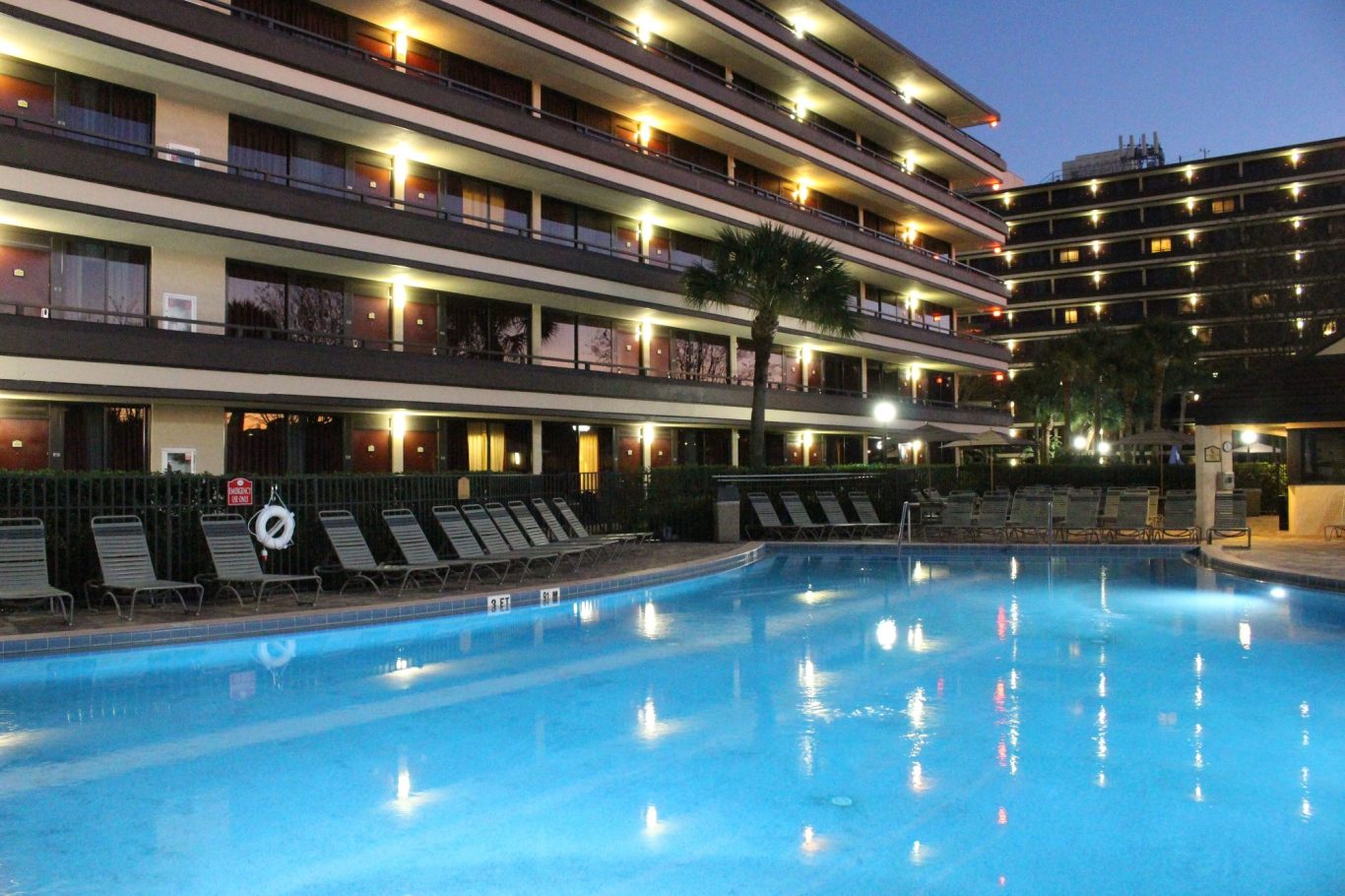 Illuminated pool area at dusk with palm trees and hotel balconies in the background.