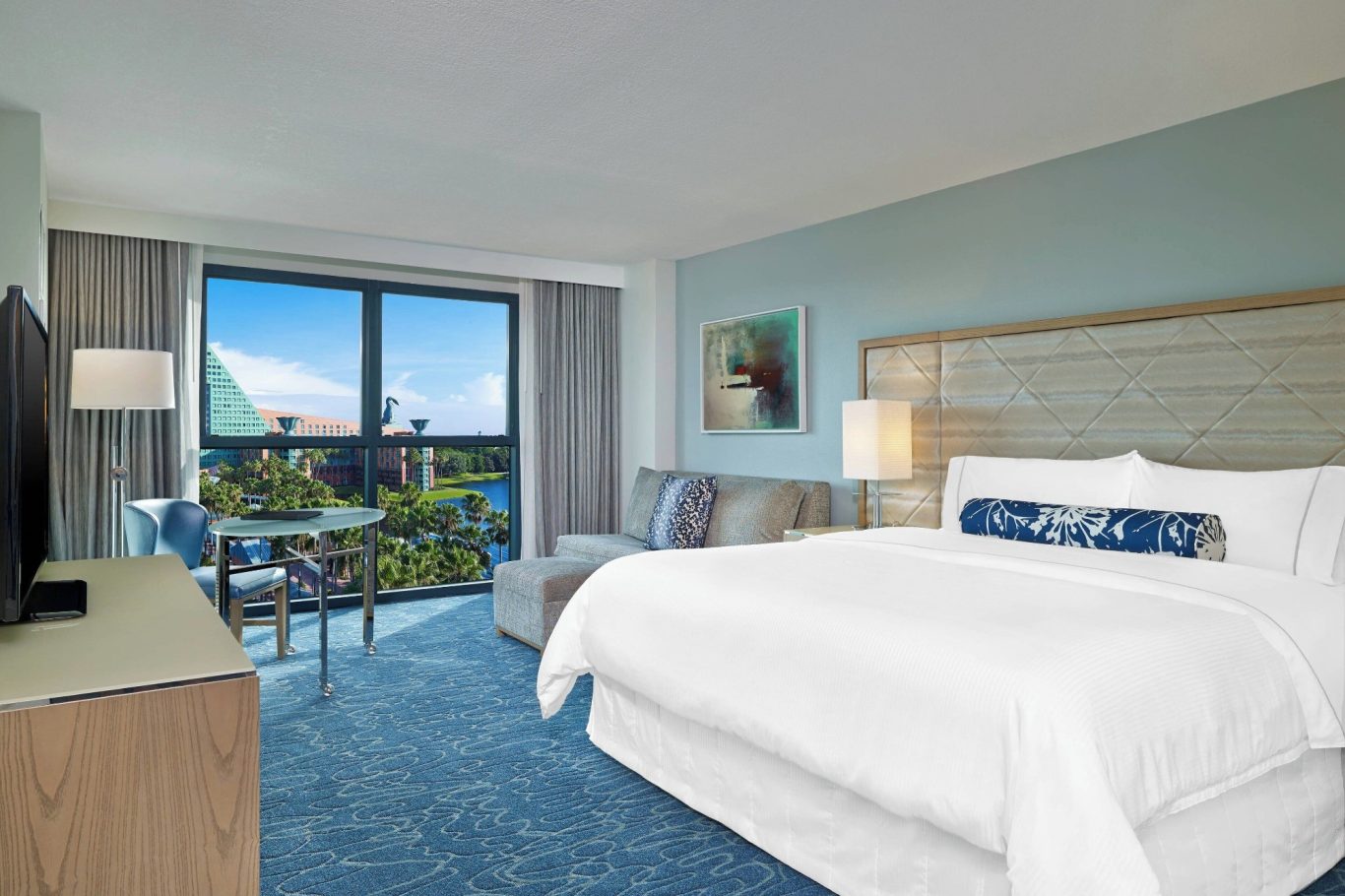 Modern hotel room with a king-size bed, a seating area, and a view of greenery outside.