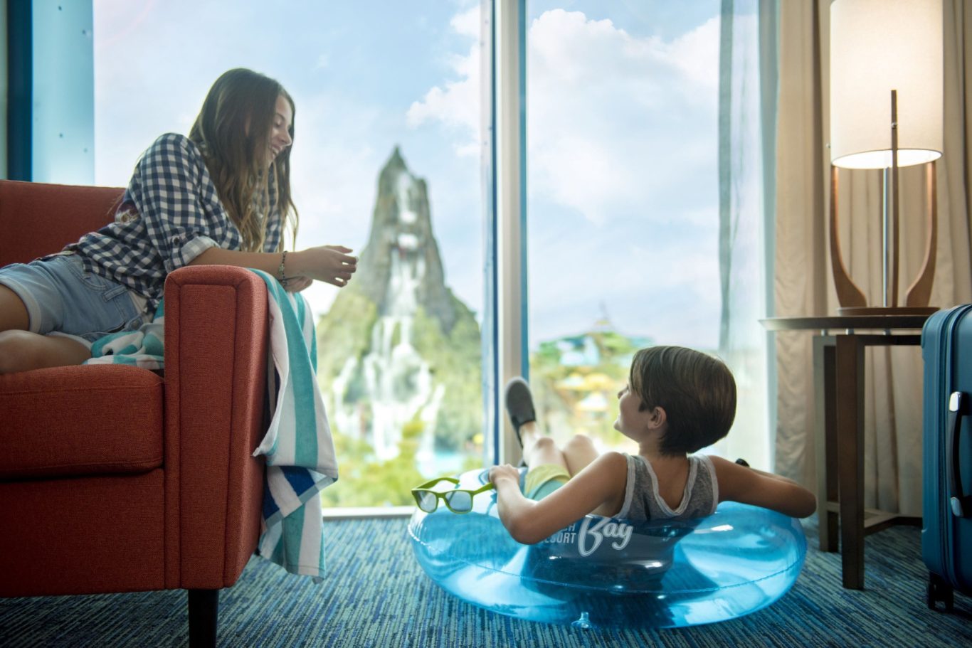 A child in a pool ring relaxes while a woman sits on a couch, with a mountain view outside.