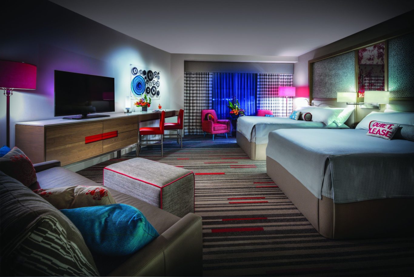 Modern hotel room with two beds, vibrant decor, seating area, and a large window.