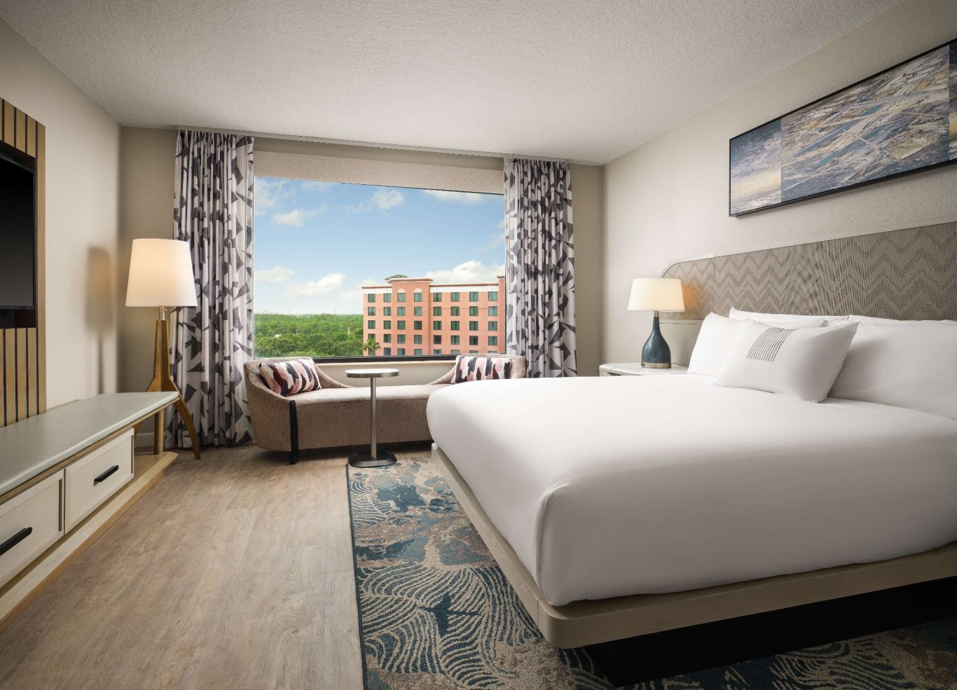 Modern hotel room with a large bed, armchairs, and a view of the outdoors.