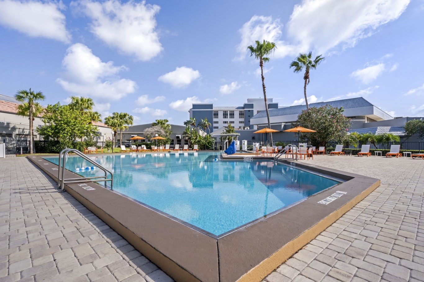 Swimming pool surrounded by palm trees and lounge chairs under a blue sky.