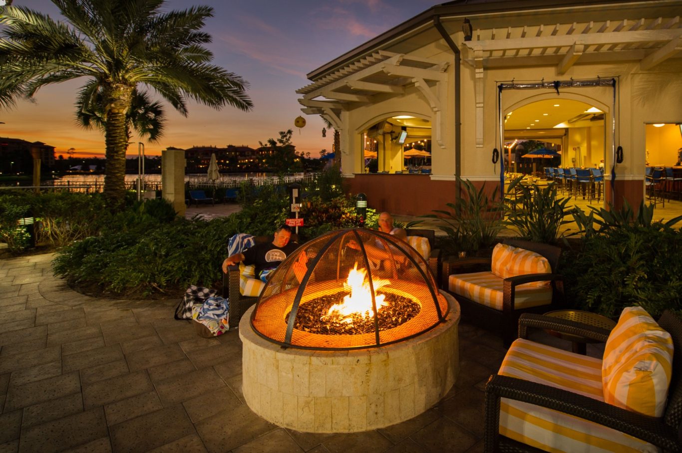 Outdoor seating area with a fire pit, surrounded by palm trees and a sunset backdrop.