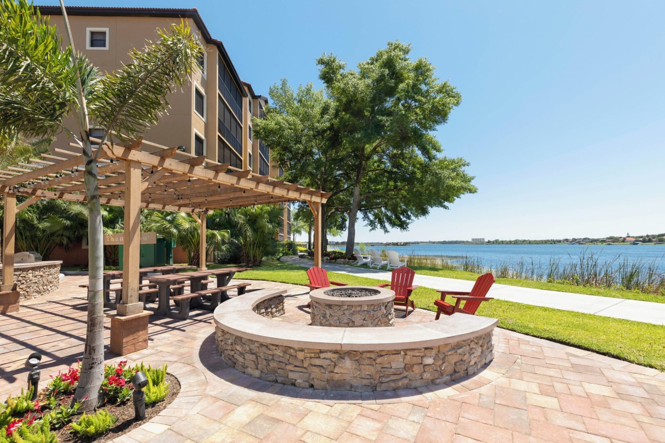 Outdoor seating area with fire pit by a lake, surrounded by greenery and a building.