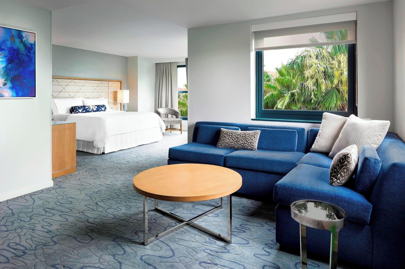 Modern hotel room featuring a blue sofa, round table, and a large window with greenery outside.