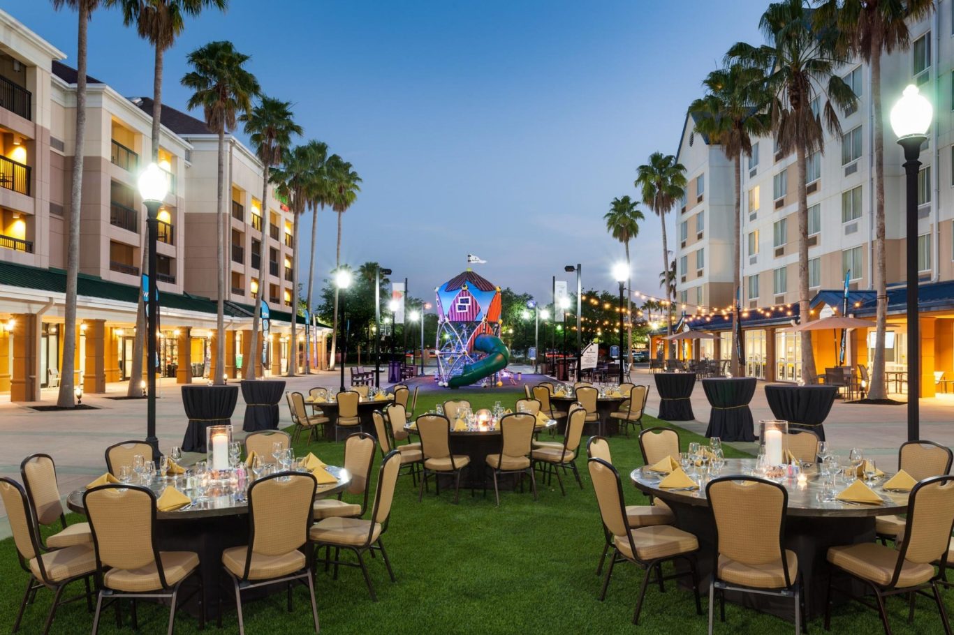 Outdoor dining setup with tables and chairs, surrounded by palm trees and buildings at dusk.
