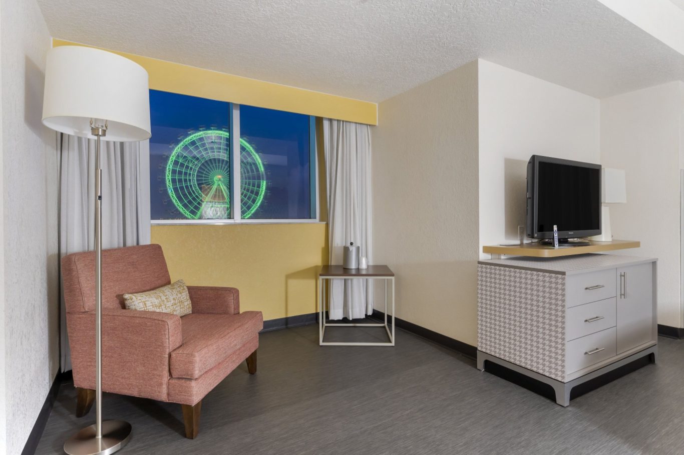 A modern hotel room featuring a pink armchair, TV, and large window with city view.
