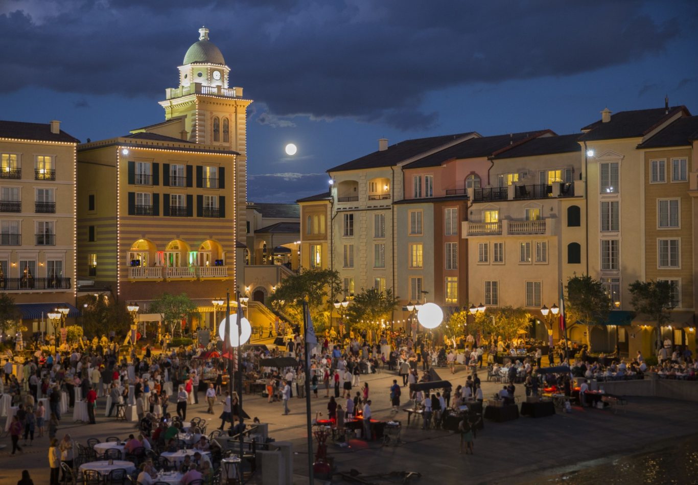 Crowded square at dusk with buildings and a large moon in the background.