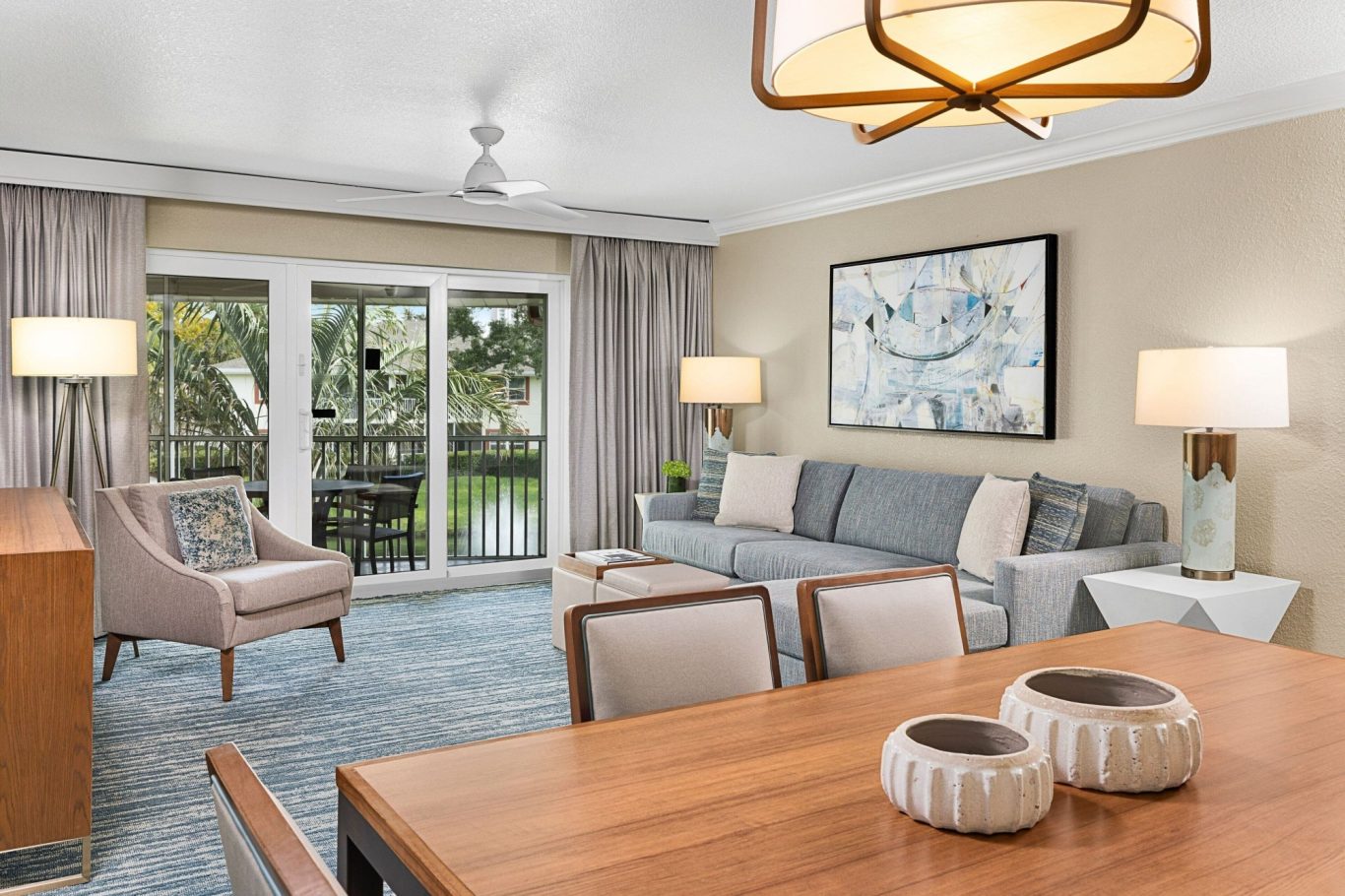 Modern living room with a grey sofa, wooden dining table, and large windows overlooking greenery.