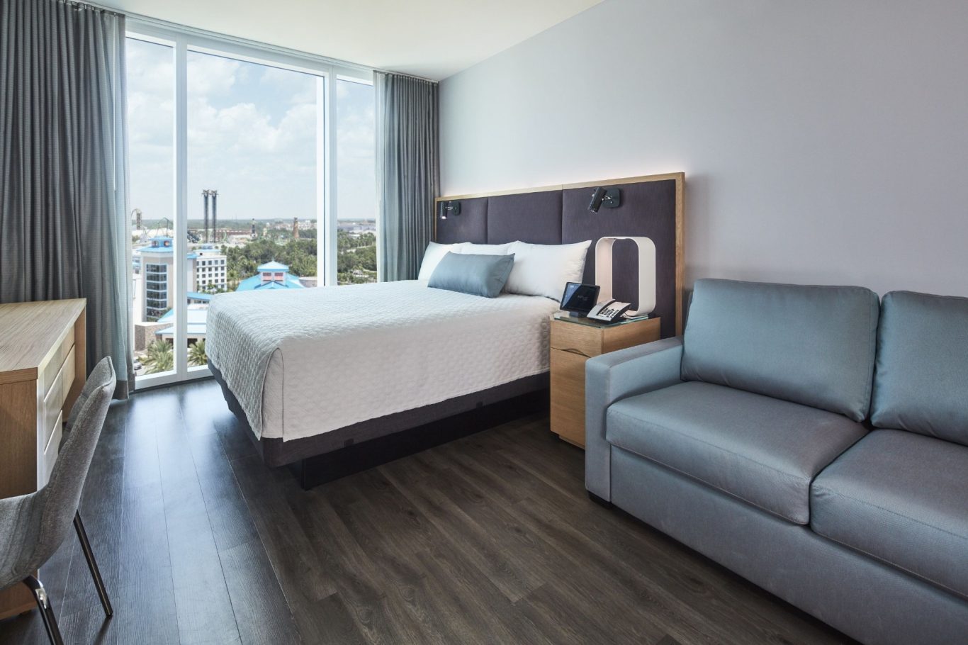Modern hotel room with a bed, desk, and seating area, large windows letting in natural light.