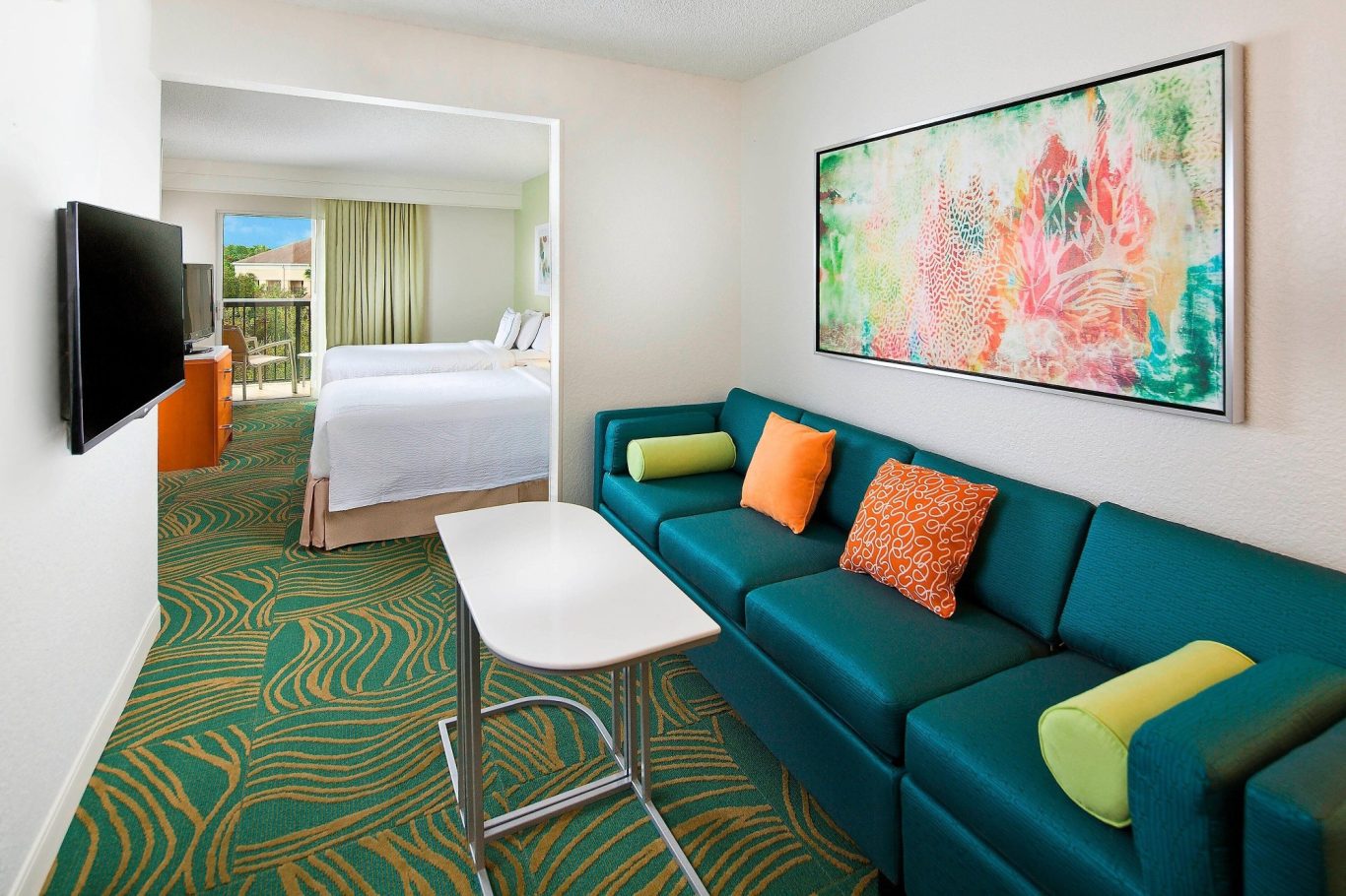 A bright hotel room featuring a teal sofa, coffee table, and vibrant artwork on the wall.