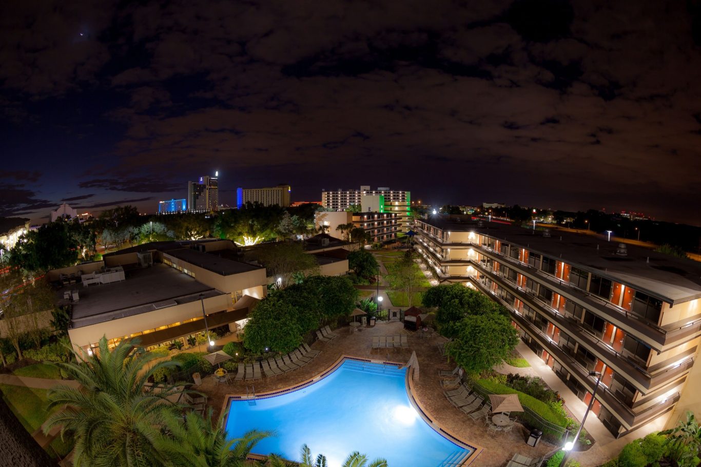 Night view of a hotel complex with a pool surrounded by greenery and city lights.
