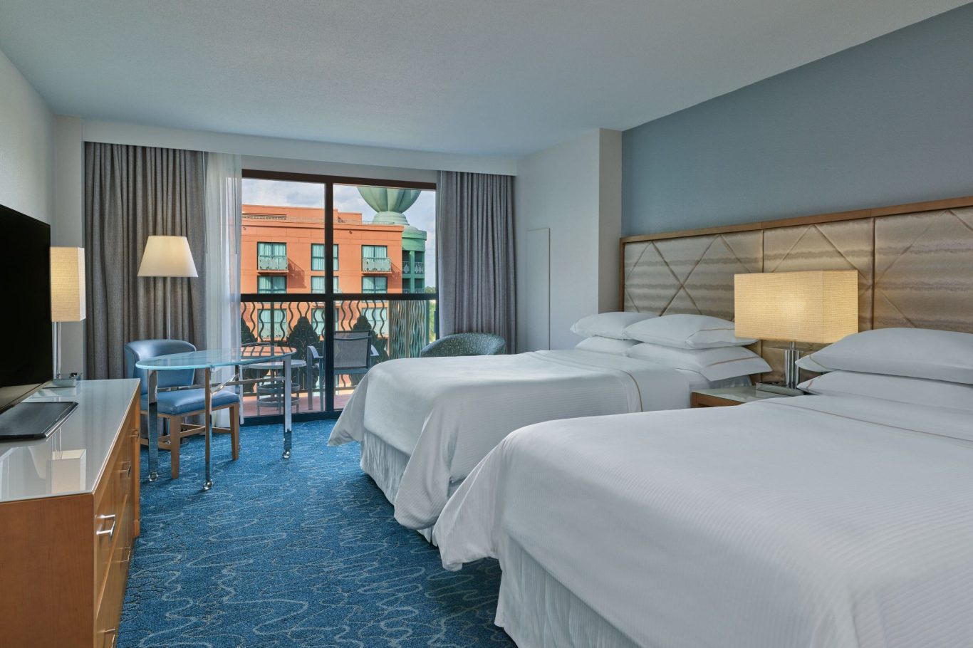 A bright hotel room featuring two beds, a TV, and a view of the outdoors.