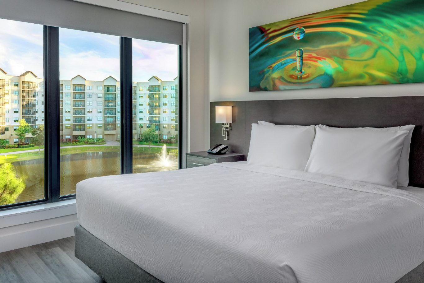 A modern hotel room with a large bed and a view of exterior buildings and greenery.