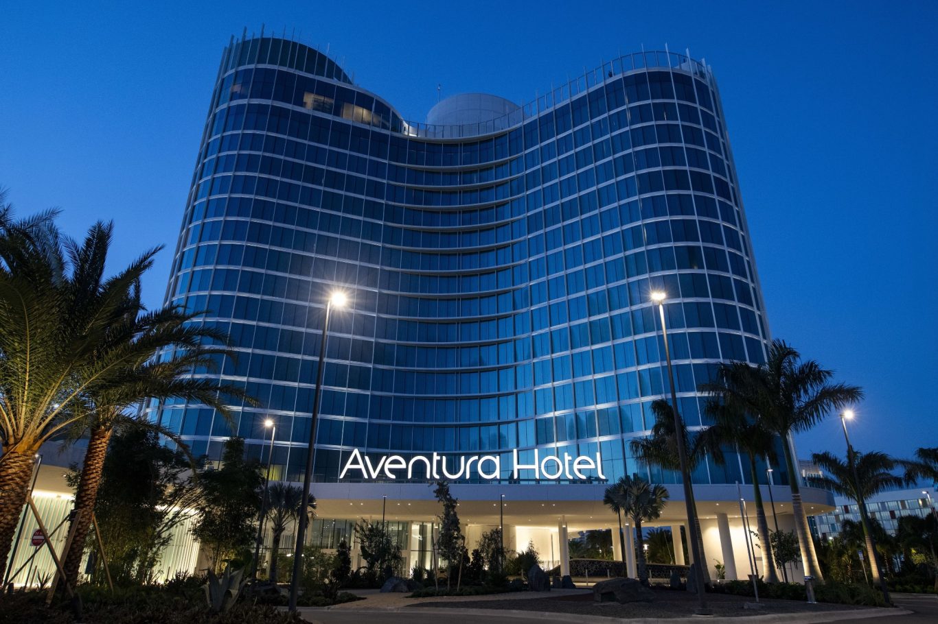 Modern hotel facade with large glass windows and palm trees at dusk.
