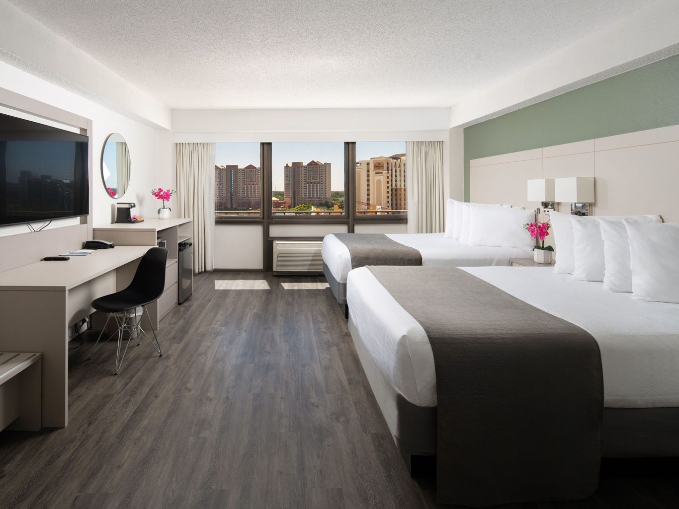 Modern hotel room with two beds, a desk, and large windows showing city views.