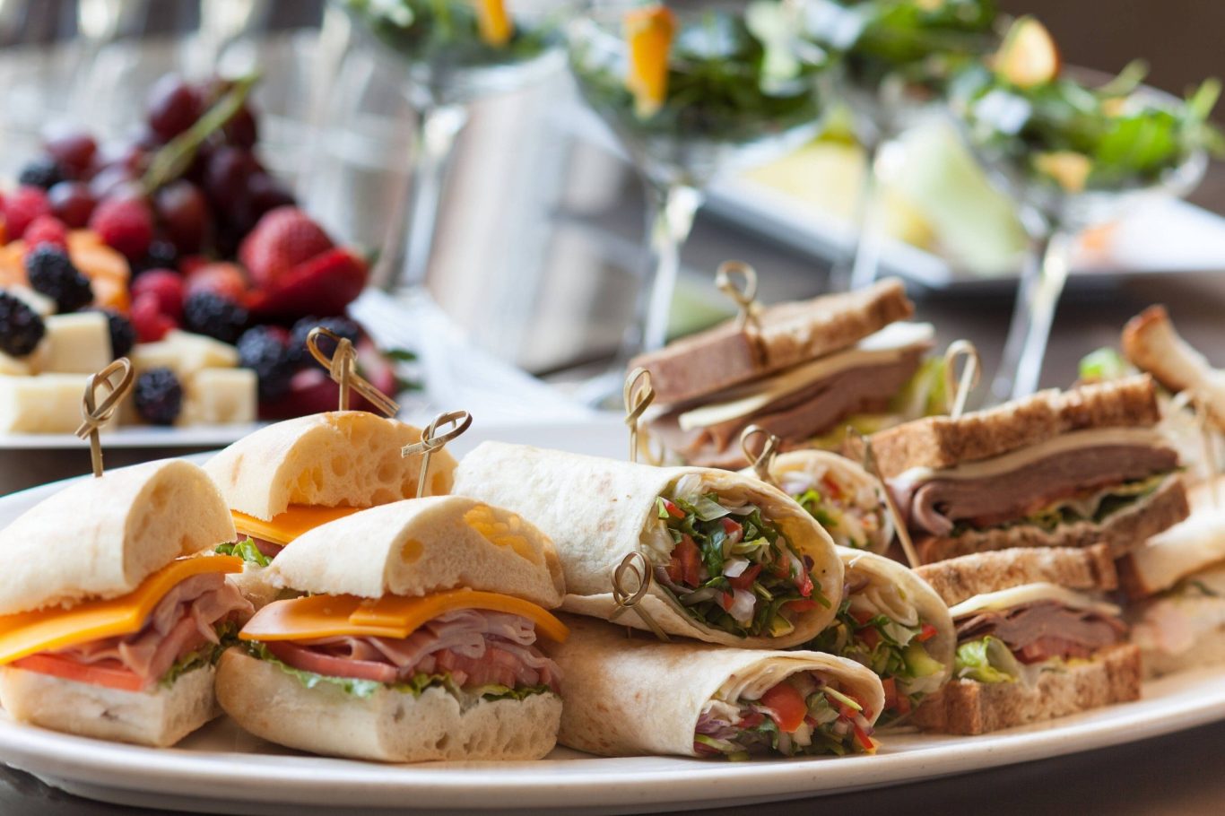 A platter of assorted sandwiches and wraps, garnished with fruit and cocktails in the background.