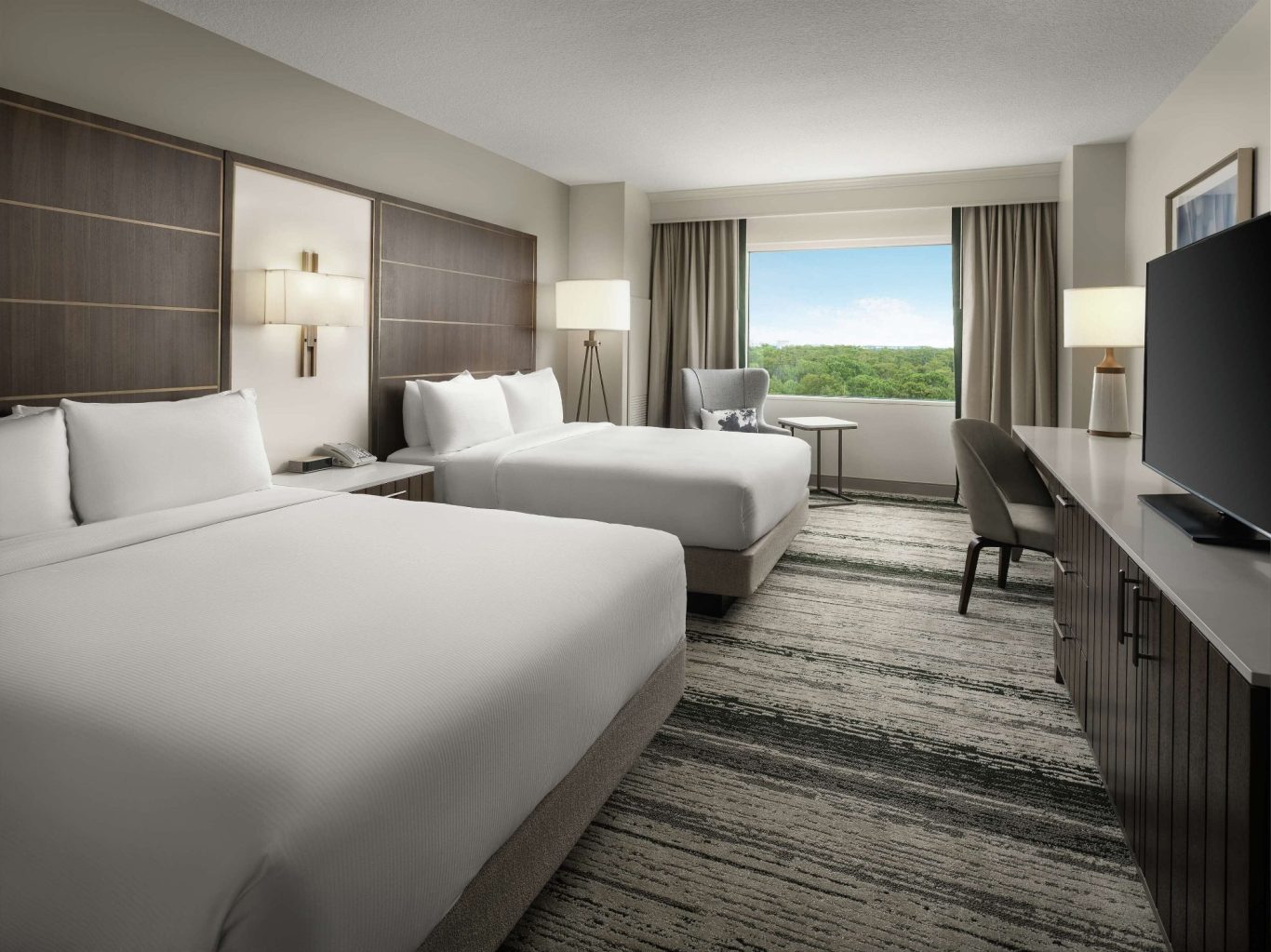 Modern hotel room with two double beds, desk, TV, and window overlooking greenery.