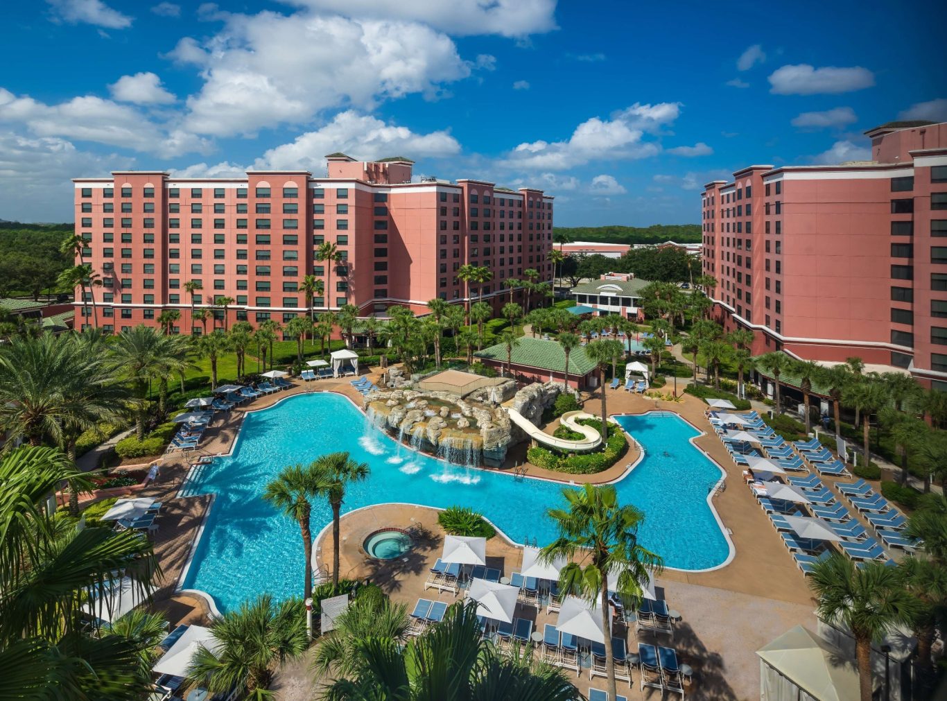 Resort with a large pool, water slide, palm trees, and luxury buildings under a sunny sky.