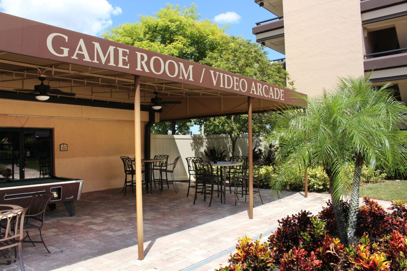 Outdoor game room with seating and signage, surrounded by tropical plants.