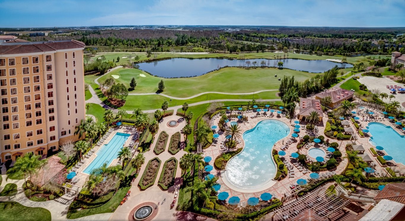 Aerial view of a resort with pools, landscaped gardens, and a lake in the background.