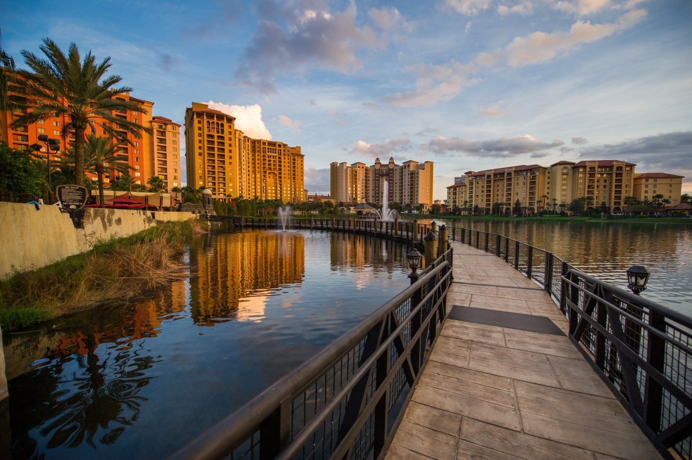 Pathway along a calm river with modern buildings and palm trees reflecting in the water.