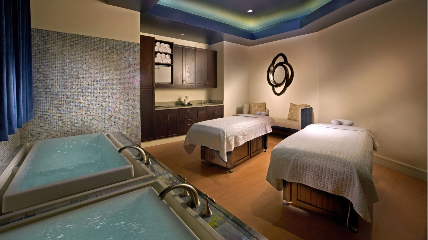 Spa treatment room with two massage tables and a soaking tub. Soft blue lighting.