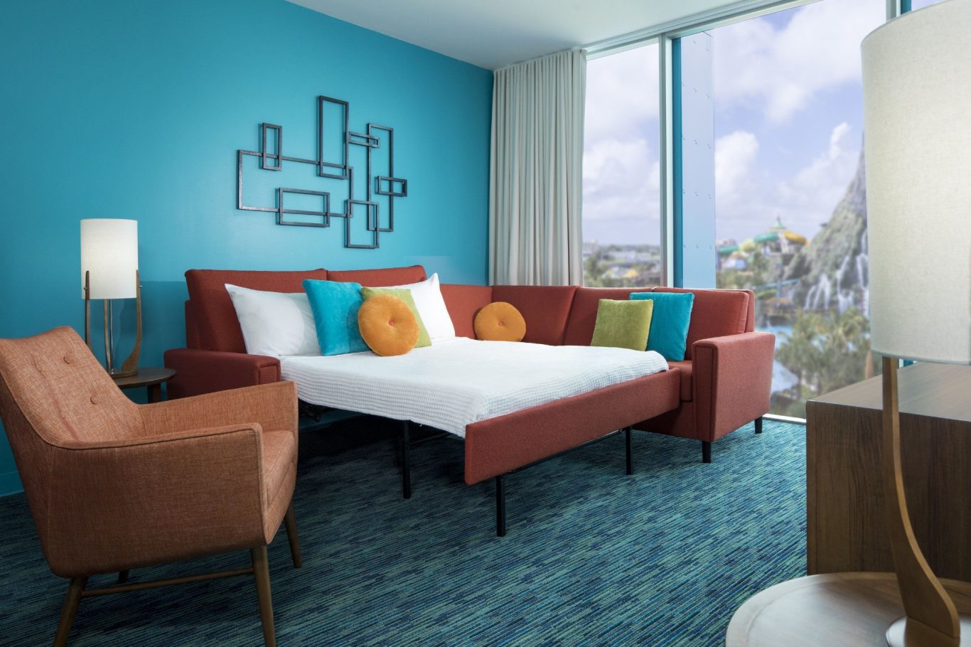 Modern hotel room featuring a turquoise wall, orange accents, a sofa, and large windows.