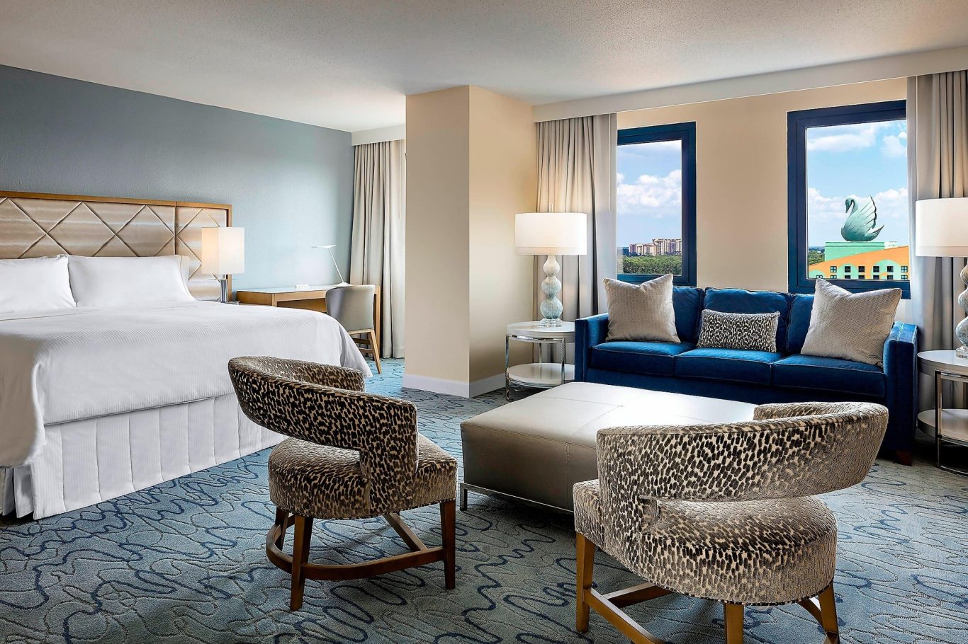 Spacious hotel room featuring a bed, sofa, chairs, and large windows with scenic views.